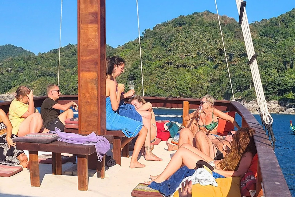 A group of people relaxing on a boat deck under a sunny sky with forested hills in the background.