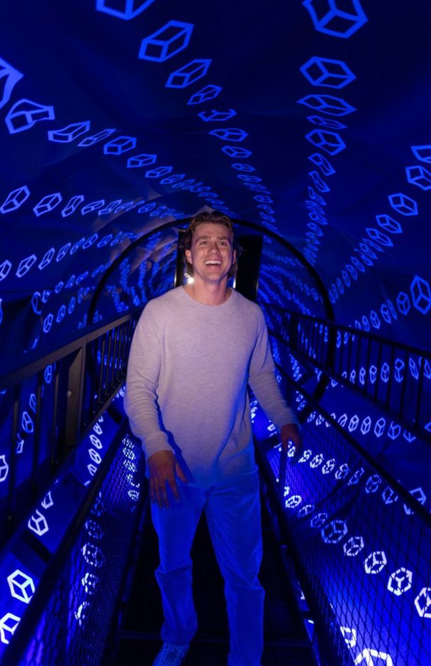 Eine Person geht durch einen mit blauen geometrischen Mustern beleuchteten Tunnel, lächelt und trägt einen hellen Pullover.