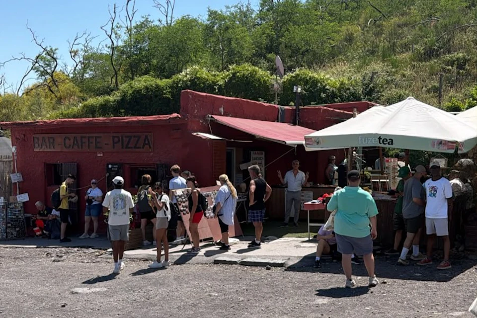 A group of people gather near a red building with a sign reading 'Pizza' and a shaded area with a counter, surrounded by greenery.