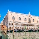 Large Gothic-style building with arched windows and ornate details, bordered by a canal with moored boats under a clear blue sky.