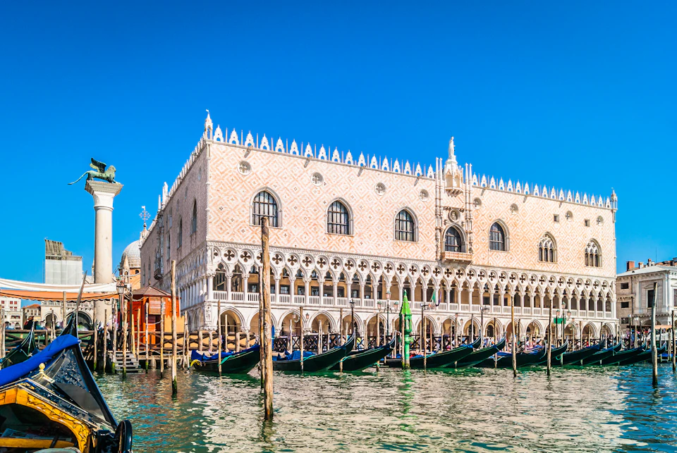 Large Gothic-style building with arched windows and ornate details, bordered by a canal with moored boats under a clear blue sky.