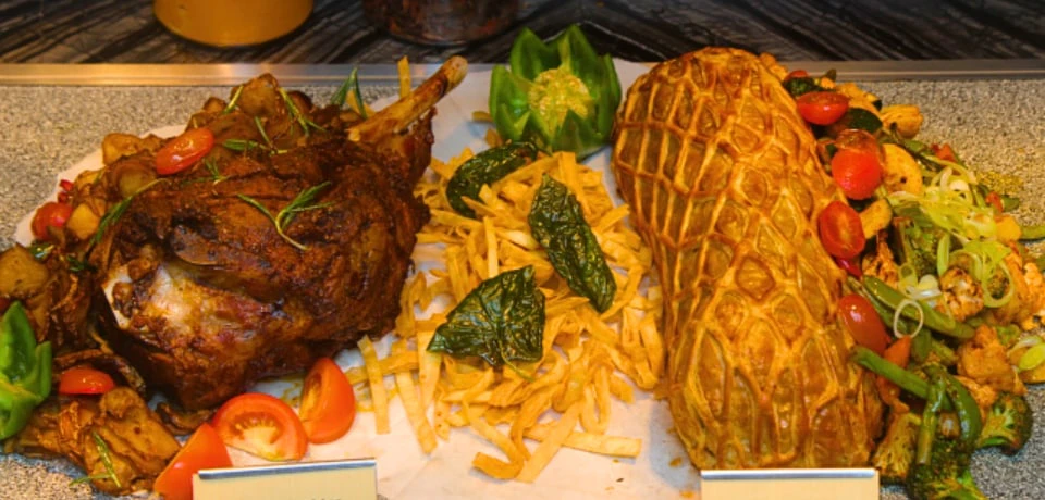 A display of cooked meat with a bone, a pastry-covered item, fries, and garnishes, including tomatoes and a carved green pepper.