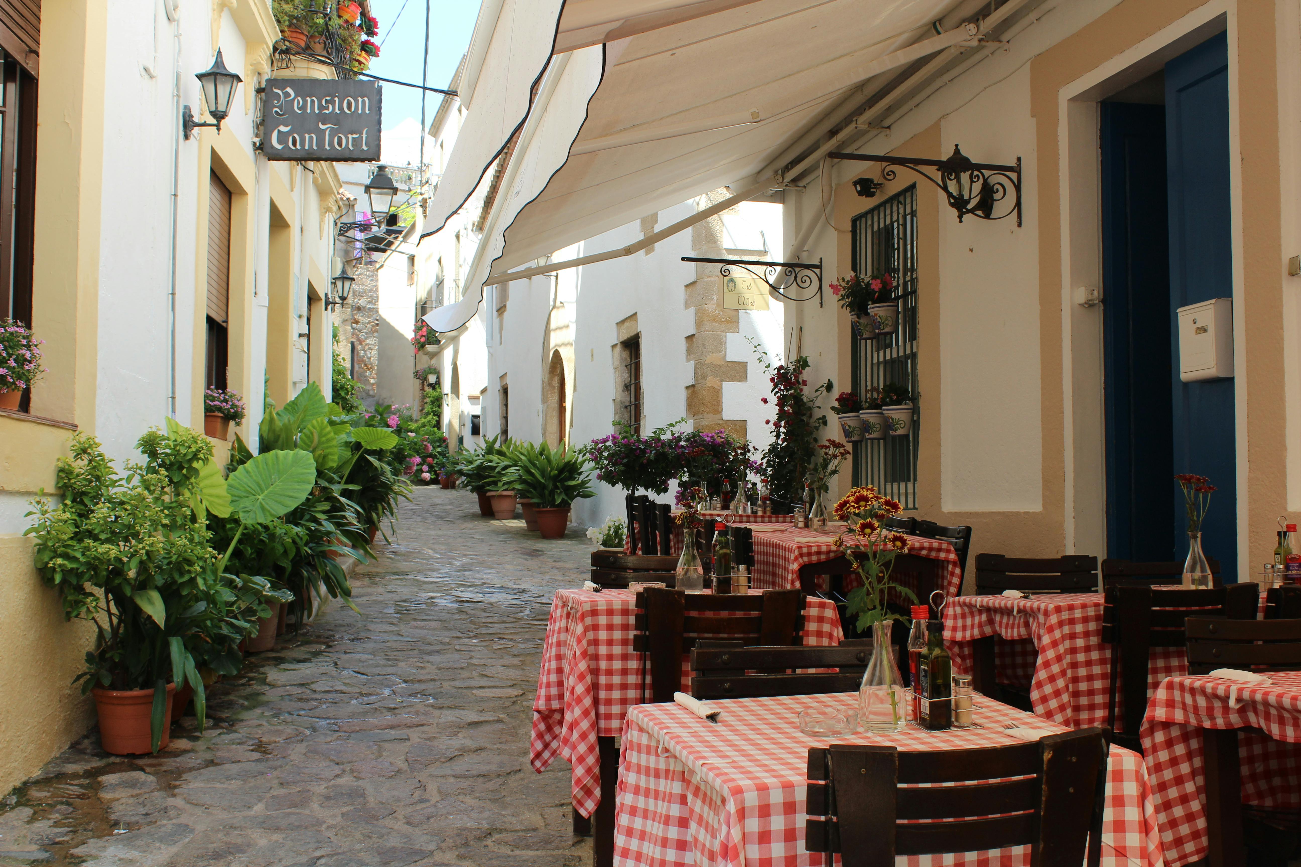 狭窄的鹅卵石小巷，带户外座椅；桌子上有红色格子桌布、植物和吊牌。