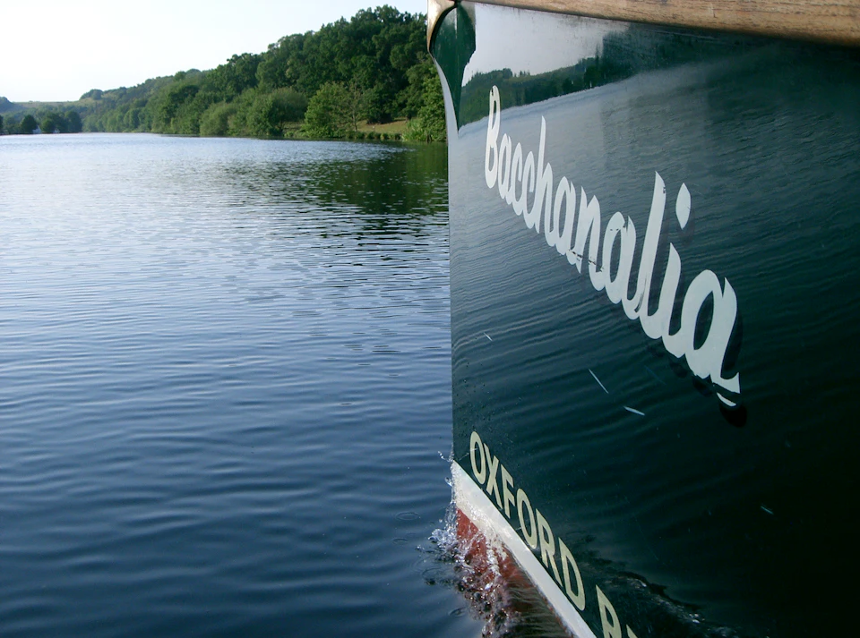 Close-up of a boat with "Bacchanalia Oxford" written on the side, surrounded by calm water and lush, green trees.