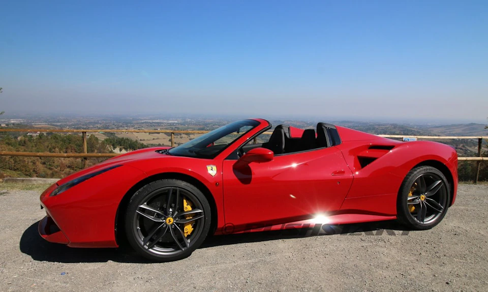 Red convertible sports car parked on a scenic overlook with distant hills and blue skies in the background.