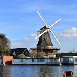 En väderkvarn står bakom husbåtar på en vattenväg under en klarblå himmel, med träd och fler hus i bakgrunden.