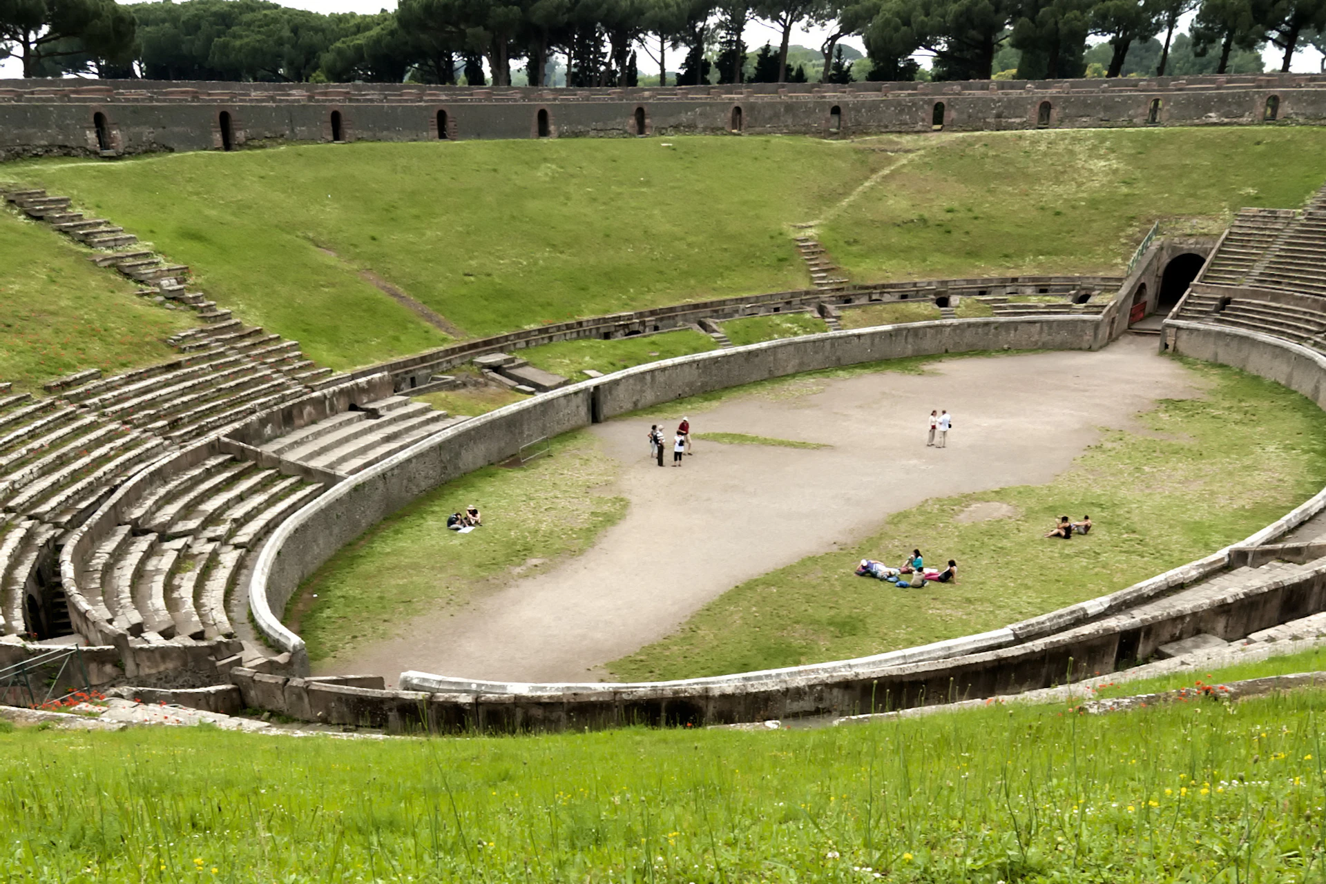 An ancient amphitheater with grass-covered seating and a central open area, with a few people walking and sitting around.