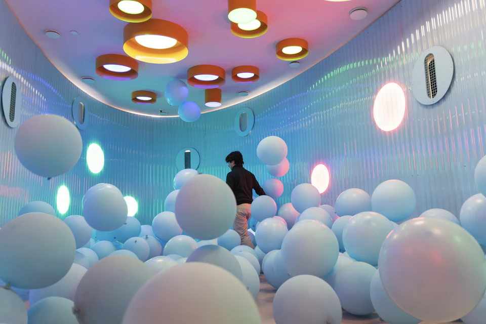 A person walking through a room filled with light blue balloons, colorful lights, and circular ceiling fixtures.