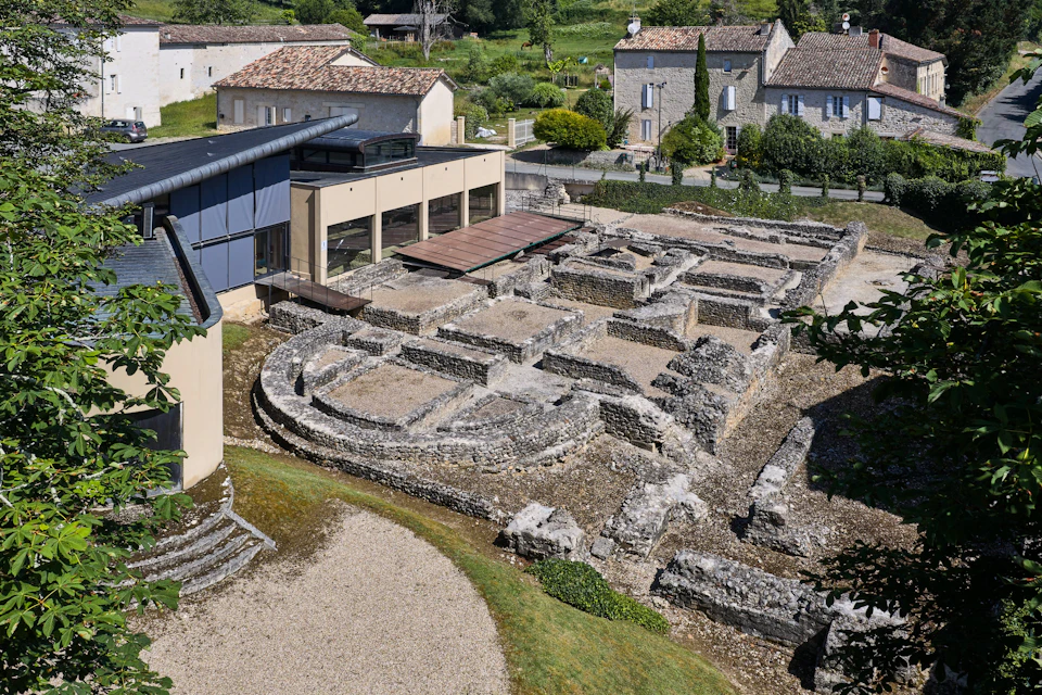 Archaeological ruins with stone foundations, surrounded by modern and traditional buildings with tiled roofs, greenery in the background.