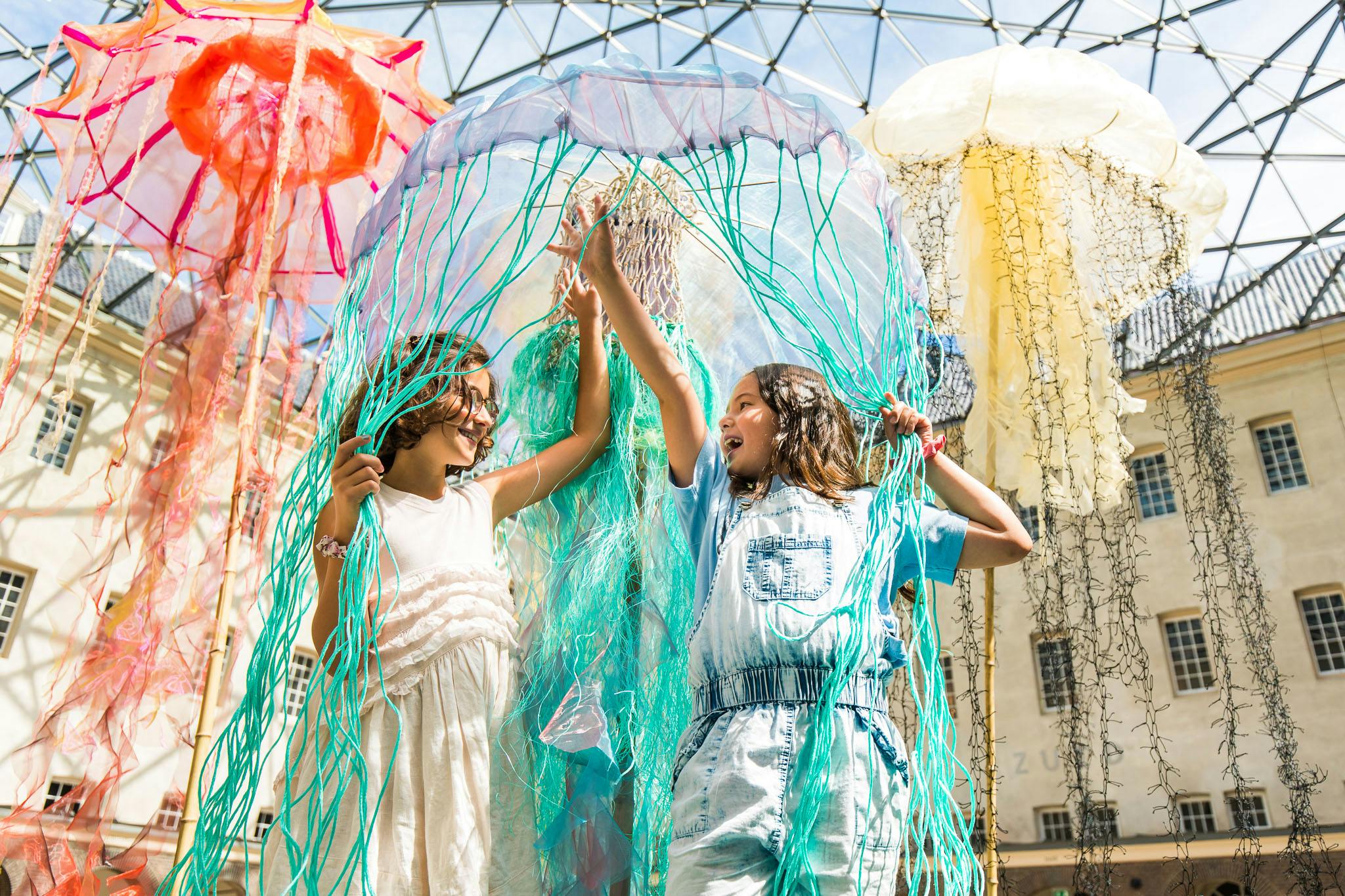 Due bambini interagiscono con gioia con grandi e colorate installazioni artistiche di meduse sotto un soffitto di vetro in un ambiente interno.