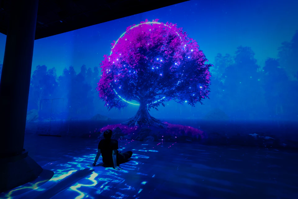 A person sits on a reflective surface under a vibrant, glowing tree with pink foliage and a ring of light in a blue-lit scene.