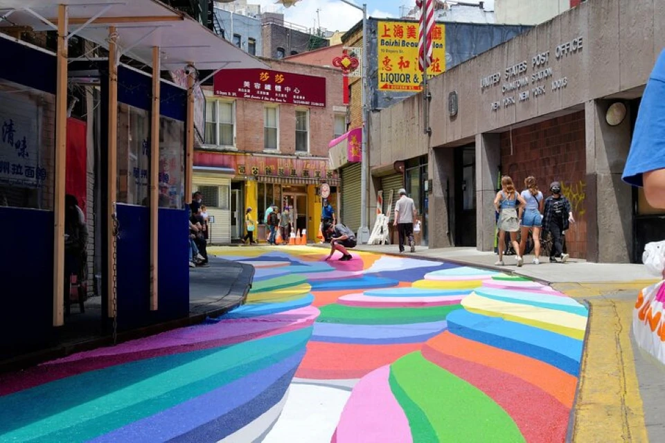 People walk past a colorful street mural near a United States Post Office in an urban area with various storefronts.