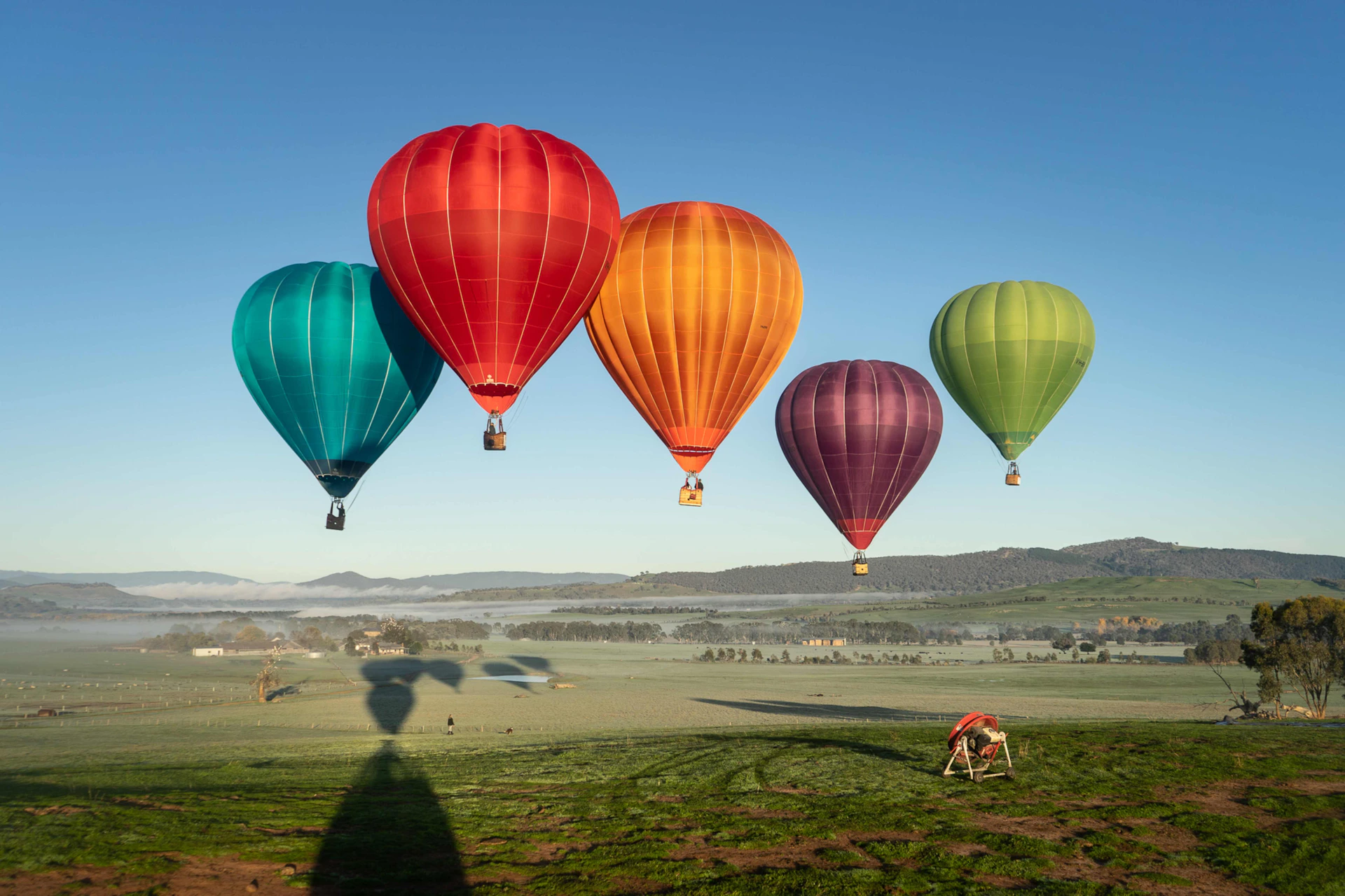 Five colorful hot air balloons floating over a green field with hills and a clear sky in the background.