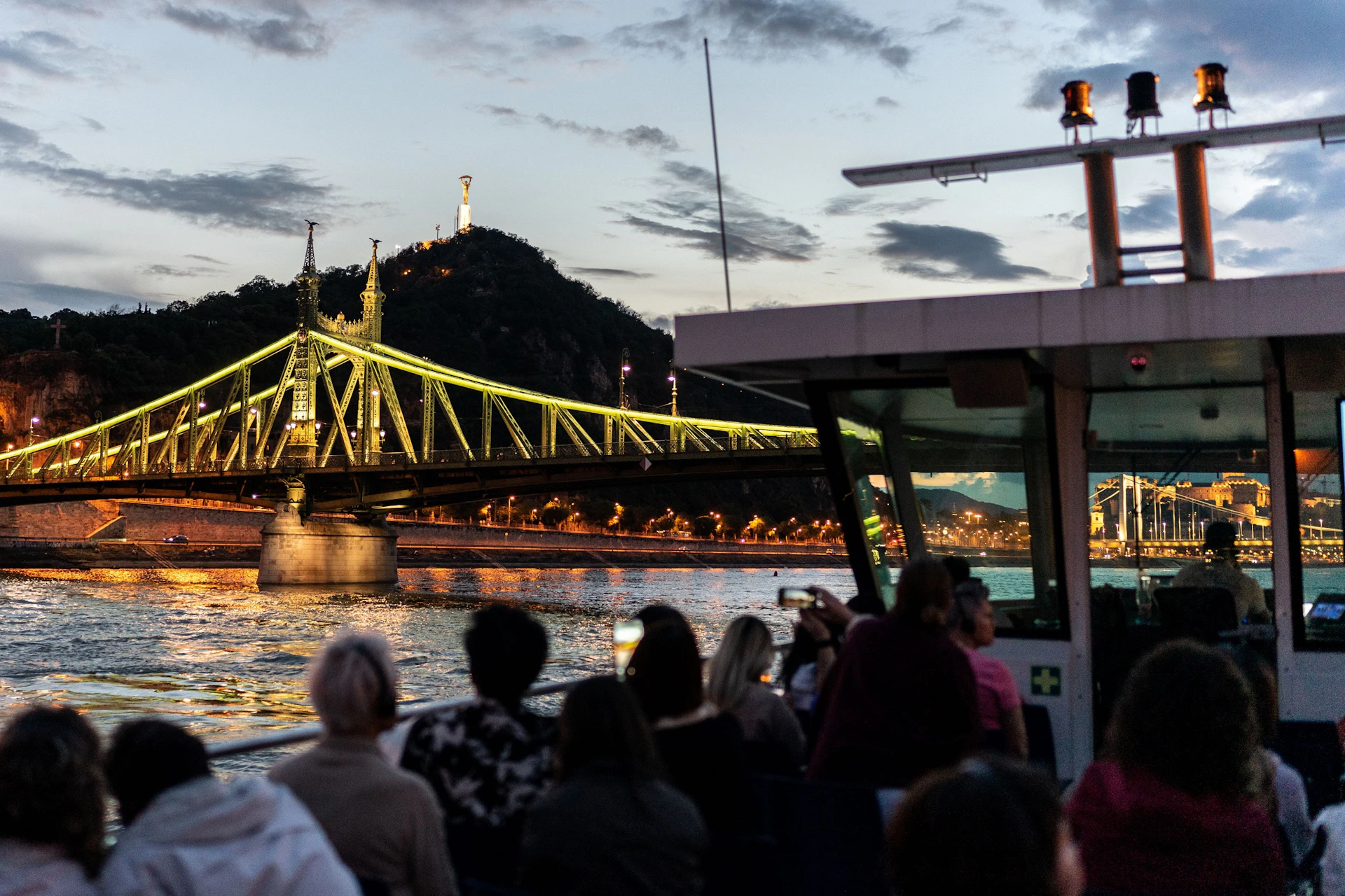 People on a boat viewing a lit bridge and a hill with a monument at dusk.