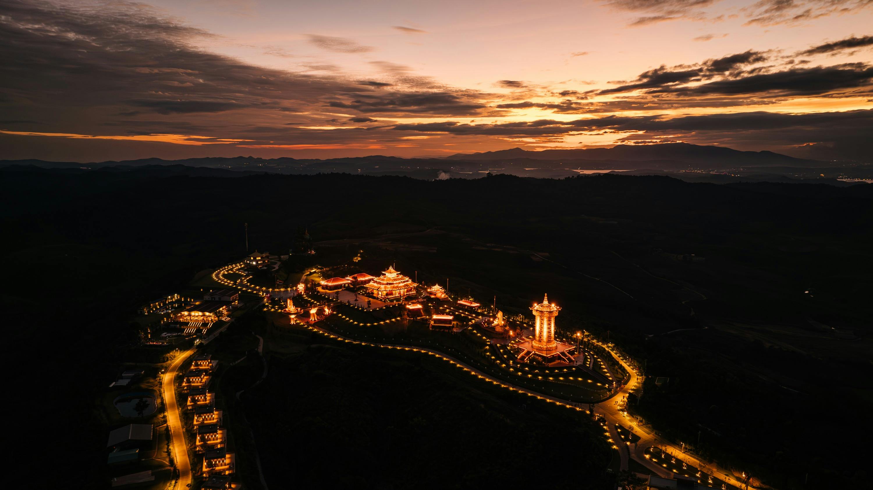Aerial view of a lit temple complex at dusk, with a glowing sky and distant hills visible in the background.