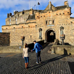 Ett par som går på en kullerstensväg mot ett gammalt, historiskt stenslott under en klarblå himmel.