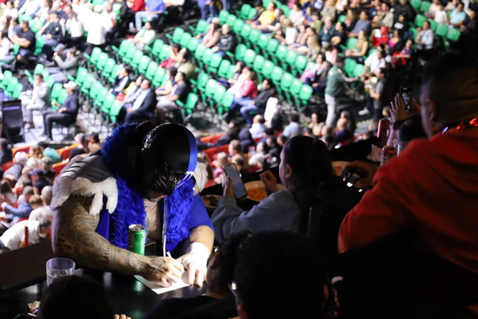 A person in a blue and black costume signs an autograph in a crowded arena with green seats.