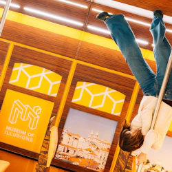 Person som hänger upp och ned på en stolpe inne i Illusionsmuseet, med gula geometriska mönster och stadsfoton på väggarna.