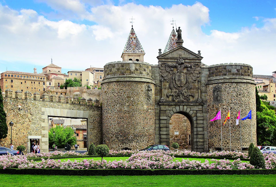 Historic stone gate with two towers, ornate crest, colorful flags, surrounded by blooming flowers and manicured lawn under a blue sky.