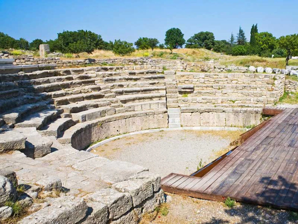 Ancient stone amphitheater with semi-circular steps, surrounded by grass, trees, and a wooden walkway leading to the center area.