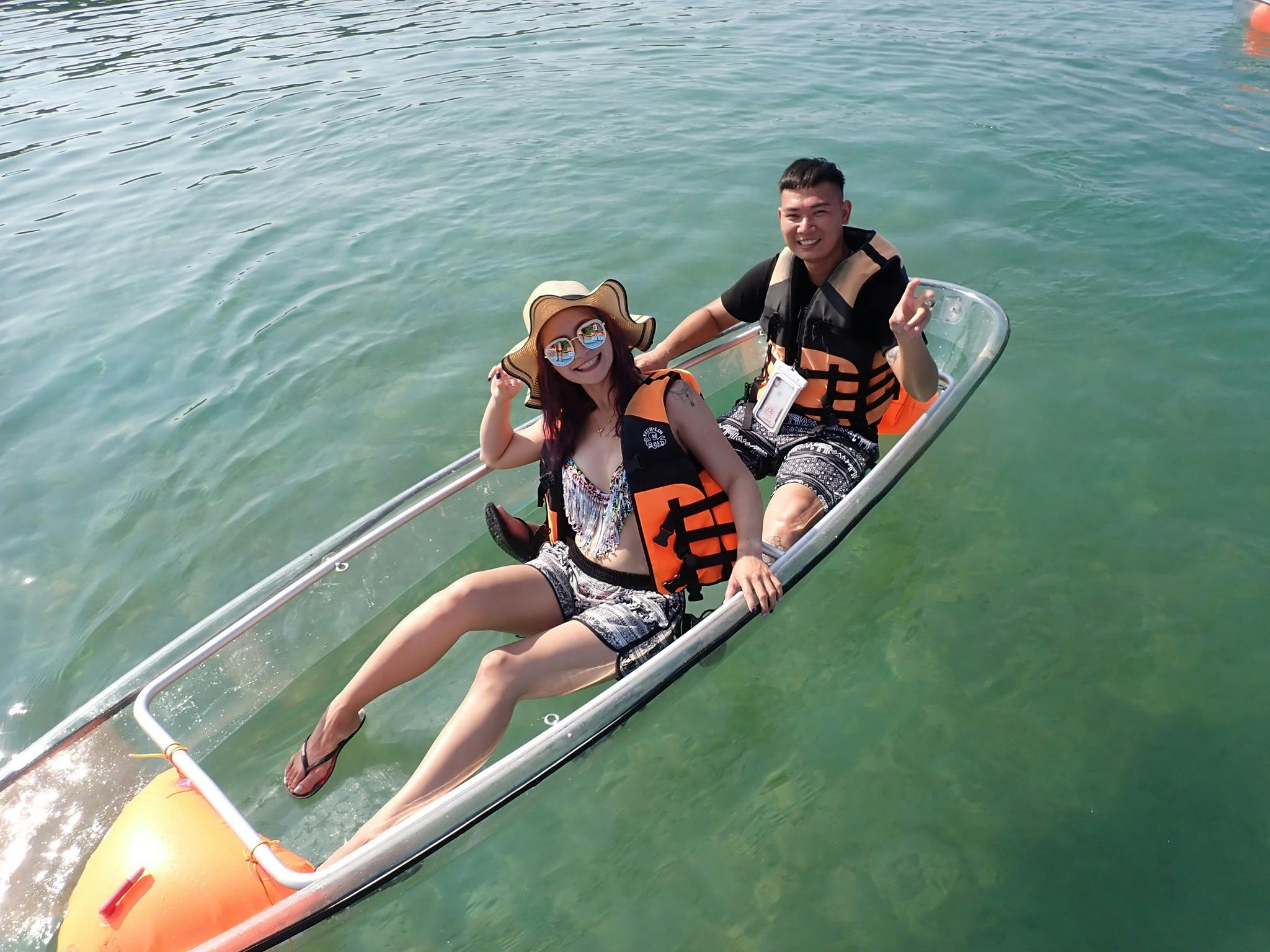 Two people wearing life jackets in a clear kayak on calm green water, one waving and the other smiling.