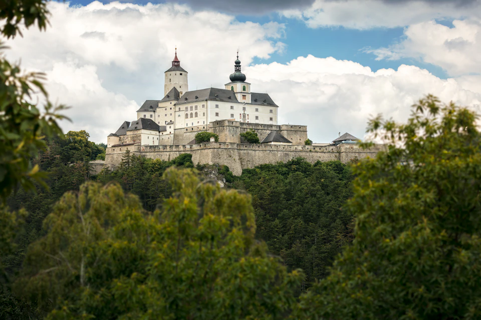 A large castle with multiple towers sits atop a hill, surrounded by dense greenery and under a partly cloudy sky.