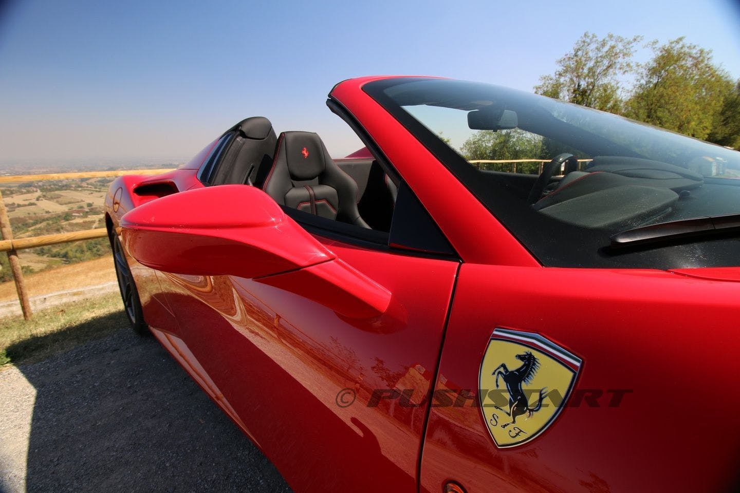 A red convertible sports car with a Ferrari logo on the door, parked on a road under a clear sky.