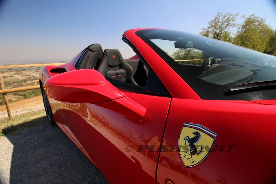 A red convertible sports car with a Ferrari logo on the door, parked on a road under a clear sky.