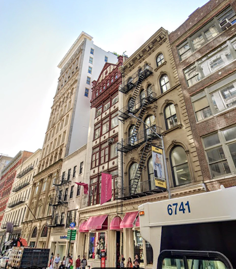 A city street lined with multi-story buildings featuring fire escapes and storefronts, with a partial view of a bus in the foreground.