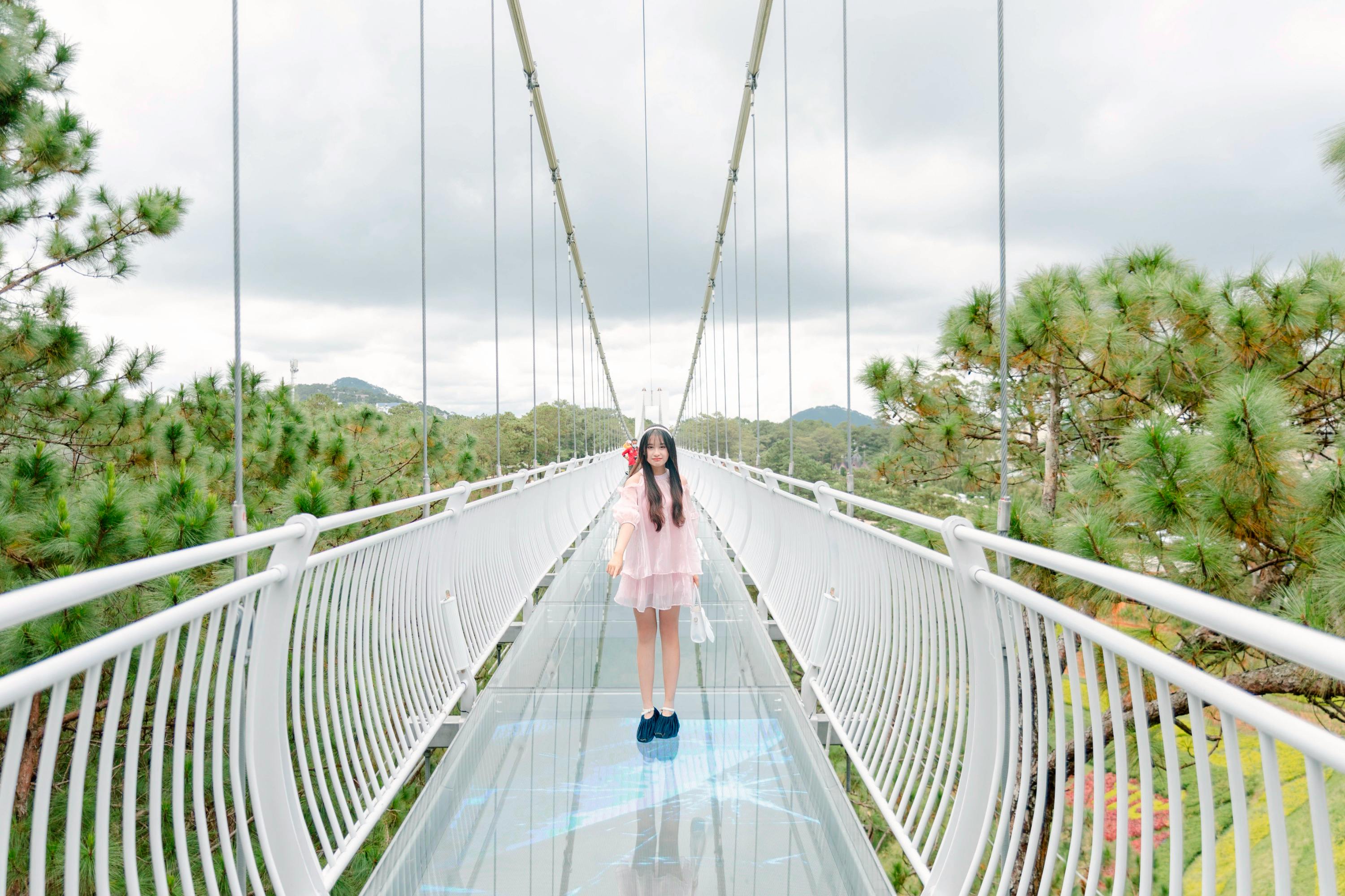 A woman stands on a glass-bottom suspension bridge surrounded by trees, with a cloudy sky in the background.