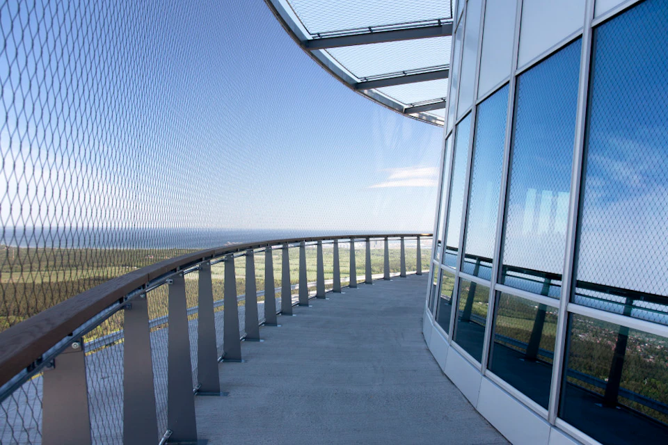 Curved walkway on a high-rise building with glass walls and a mesh safety barrier overlooking a distant landscape.