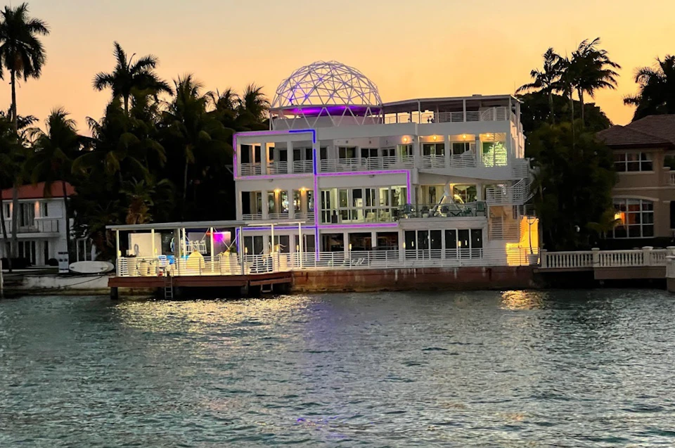 A large modern waterfront house with an illuminated dome structure on the roof, surrounded by tropical trees at sunset.