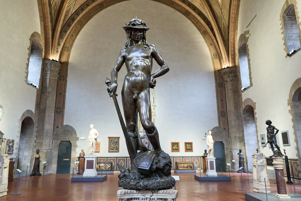 A bronze statue of David by Donatello stands in a museum hall with high arched ceilings and surrounding artwork.