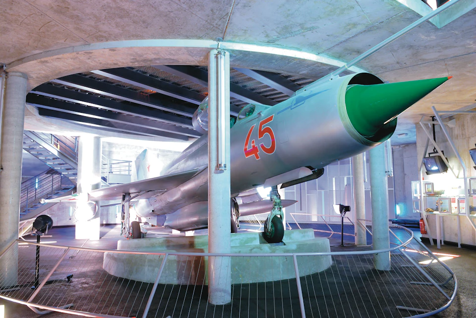 A silver fighter jet with a green nose cone and the number 45 displayed, exhibited in an indoor museum behind railings.