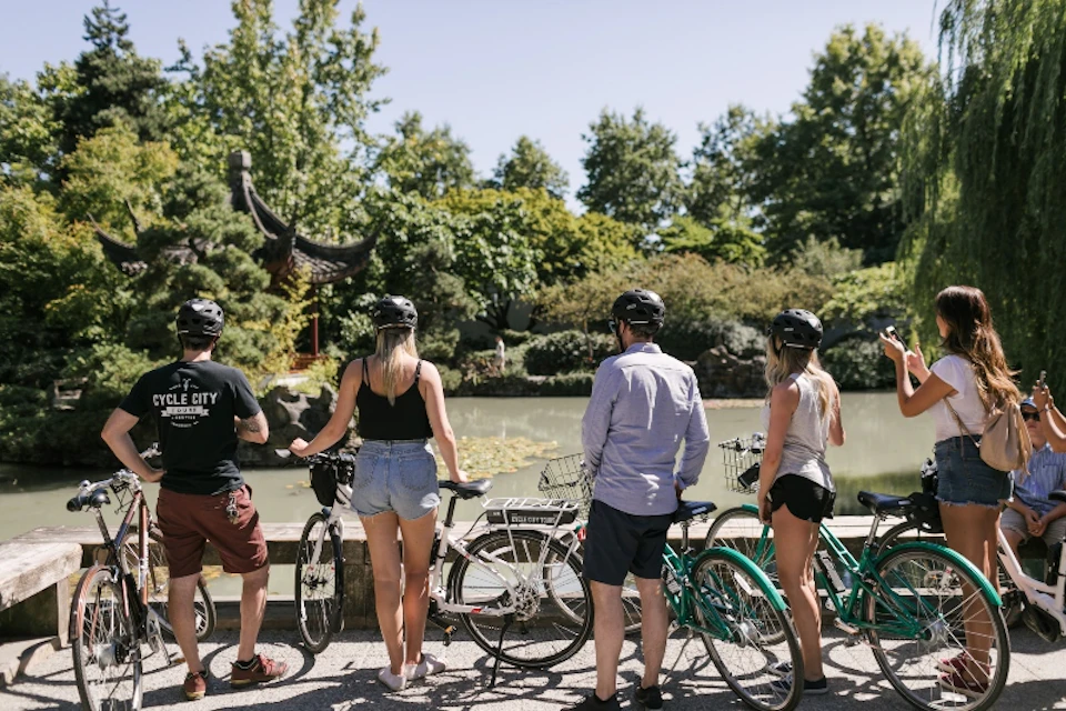 Four people with bicycles, wearing helmets, stand facing a pond with greenery and a traditional structure in the background.