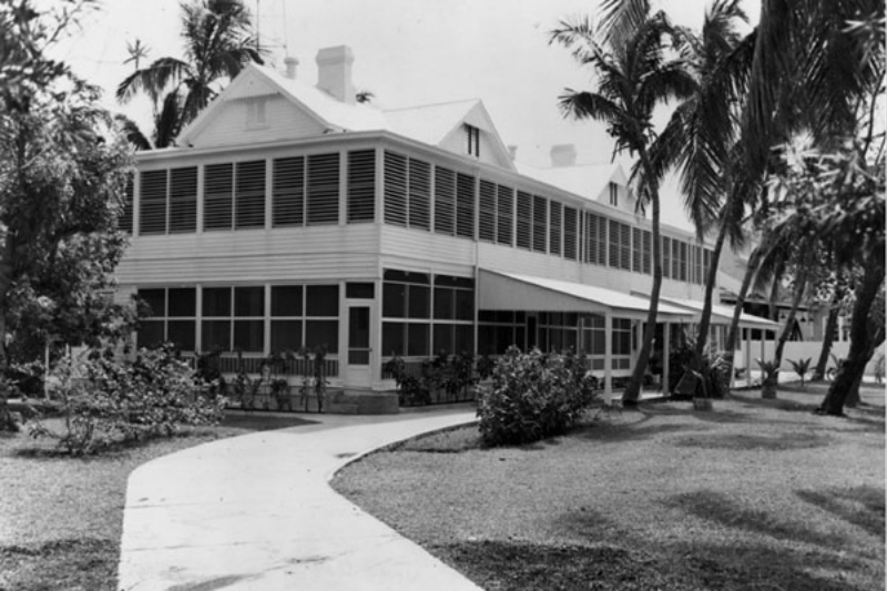 A black-and-white photo of a large, two-story building with many windows, a wrap-around porch, palm trees, and a curved sidewalk.