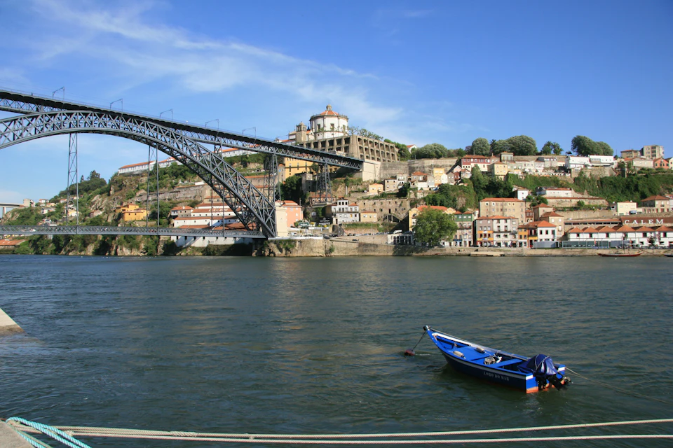 A blue boat floats on a river near a large iron bridge, with a hill of colorful buildings and a church in the background.