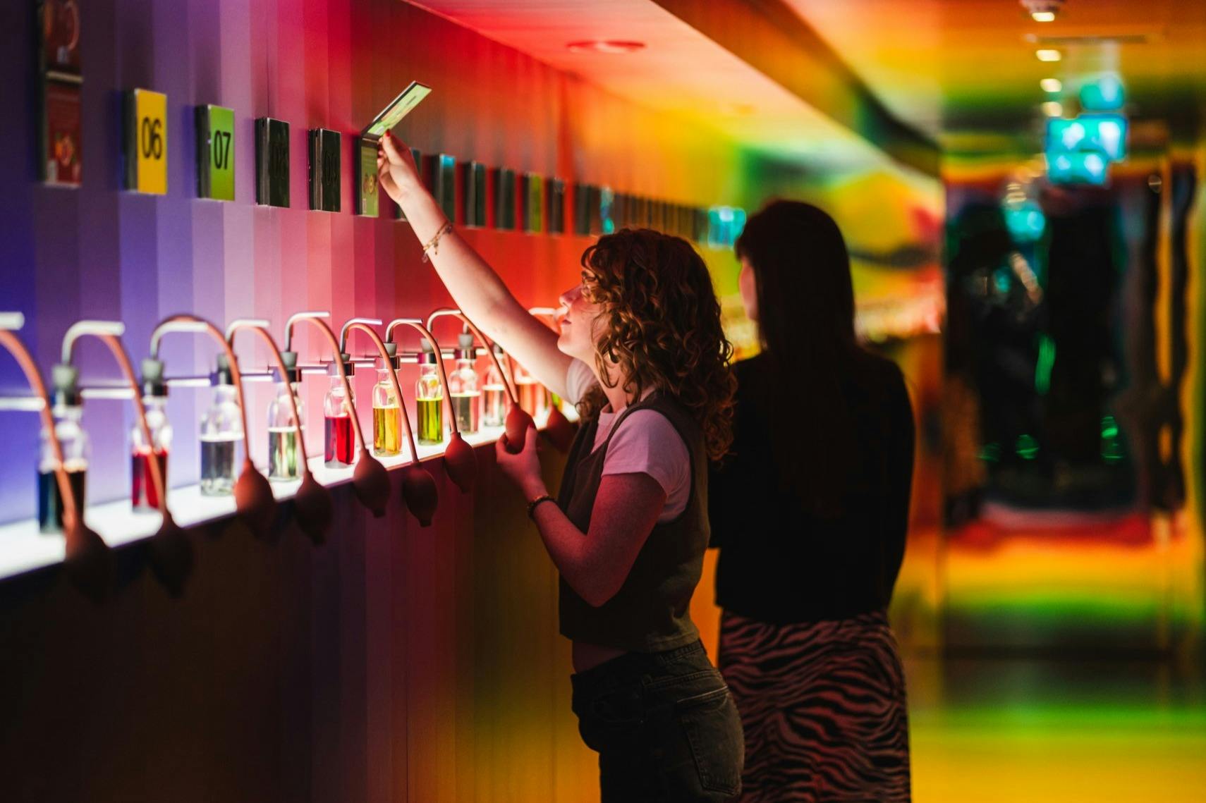 一个人在彩虹灯照亮的墙上伸手拿一张卡片，下面有小瓶子，而另一个人站在附近。