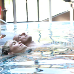 Två personer slappnar av och flyter på rygg i en inomhuspool med naturligt ljus som kommer genom stora fönster.