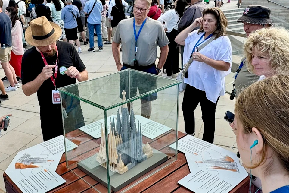 A group of people gathers around a display case containing a model of a cathedral, with a large ornate building in the background.