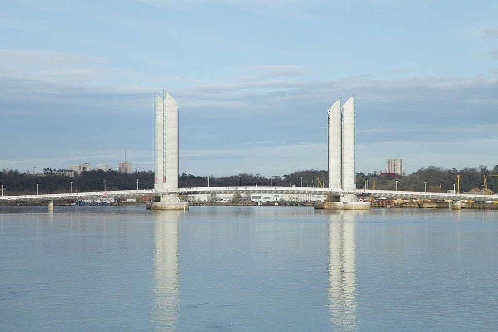 A modern drawbridge spans a wide river, with two tall central towers reflecting in the calm water under a cloudy sky.