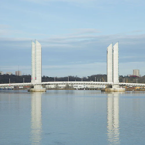 En modern vindbrygga sträcker sig över en bred flod, med två höga centrala torn som speglar sig i det lugna vattnet under en molnig himmel.