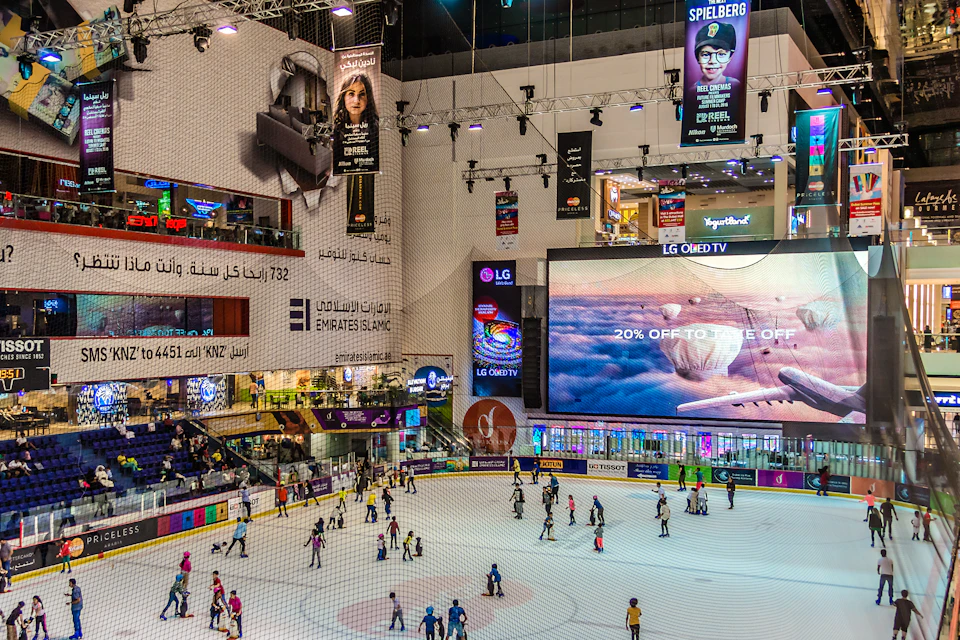 People ice skating in a large indoor rink with advertisements, banners, and a digital billboard promoting a 20% discount.