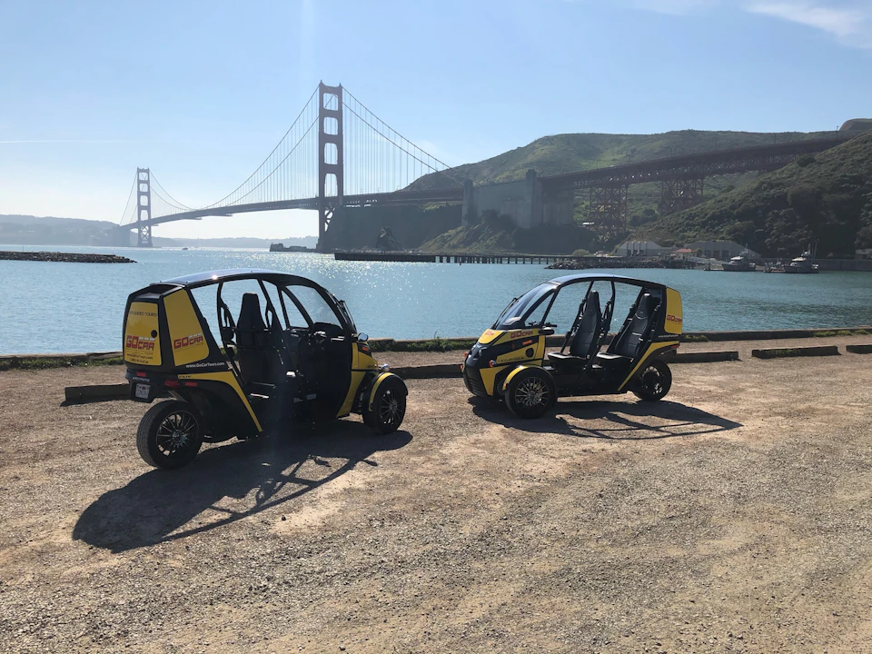 Two compact yellow and black electric vehicles parked near water with the Golden Gate Bridge visible in the background.
