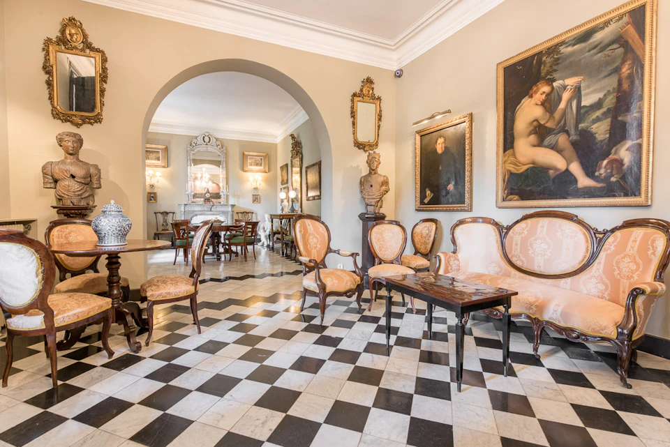 Elegant room with checkered floor, antique furniture, framed paintings, a large mirror, and a sculpture. Archway leads to another space.