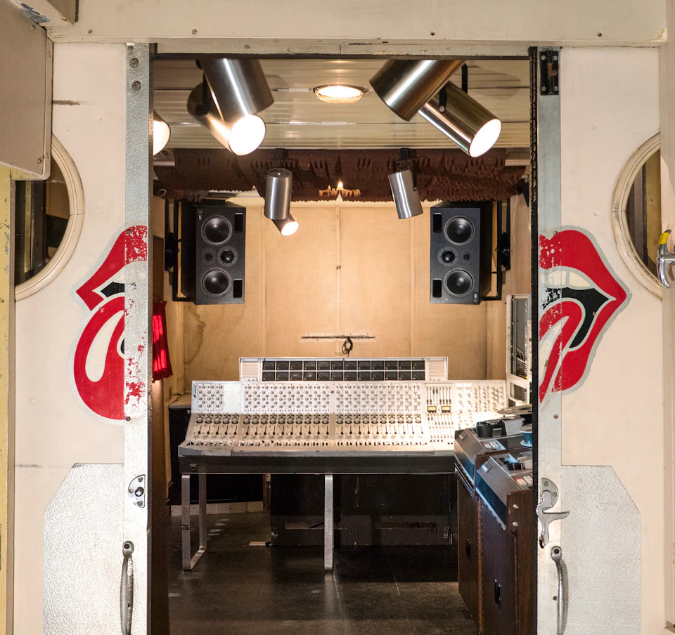A recording studio featuring a mixing console, speakers, and overhead lights, with painted red lips logos on the doors.