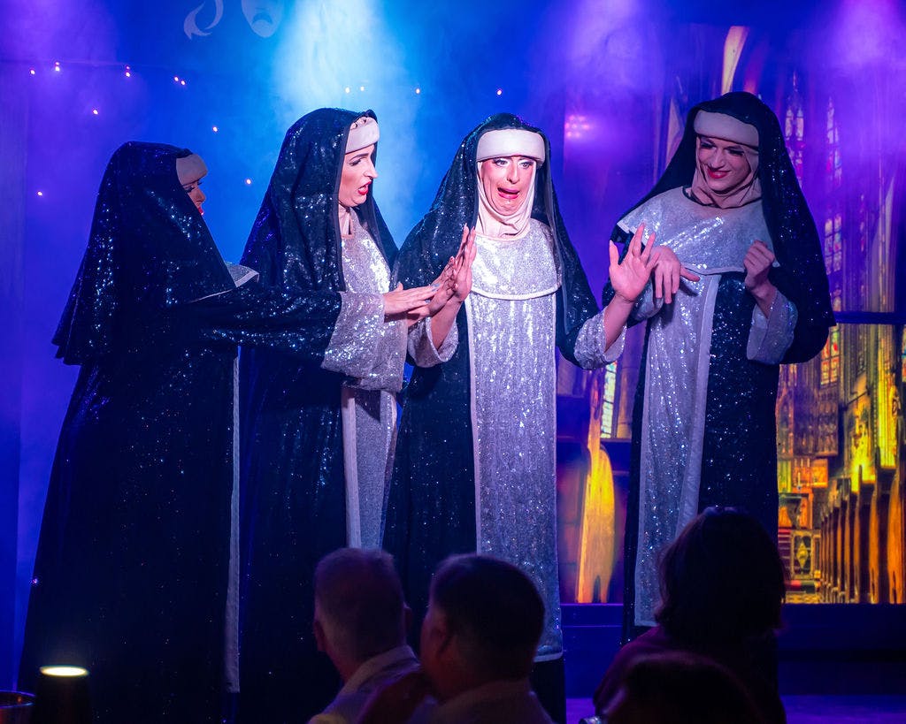 Actrices vestidas de monjas con trajes brillantes están en el escenario bajo una iluminación espectacular, con un público visible en primer plano.