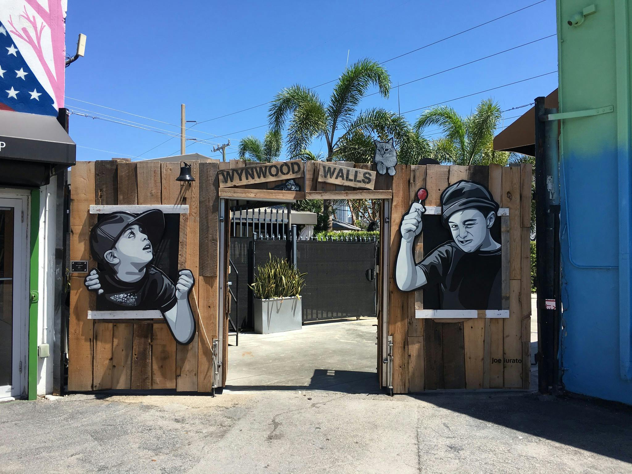 Entrance to Wynwood Walls featuring murals of a boy peeking through a wooden fence and another boy holding a lollipop, under a clear sky.