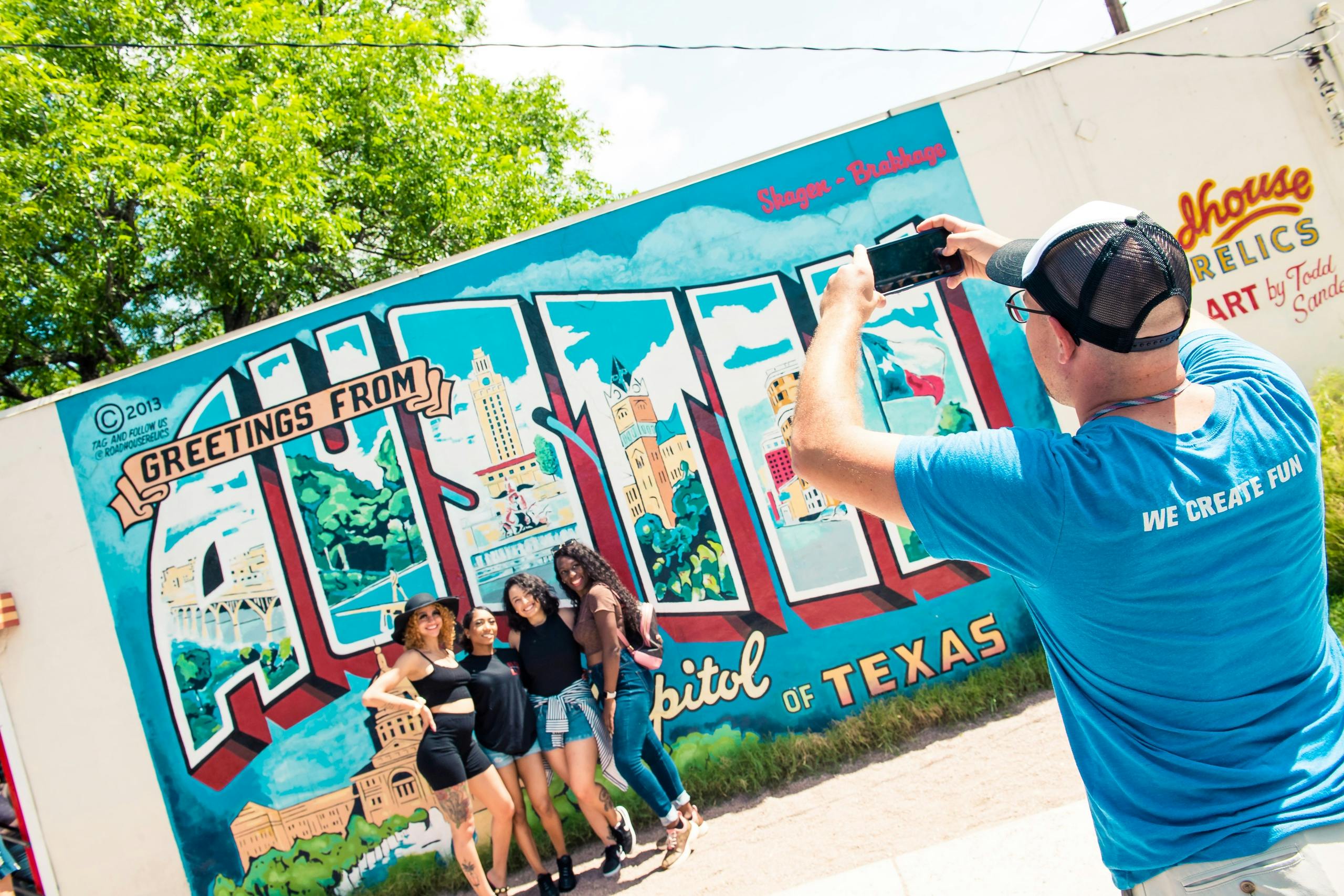 Un hombre con camisa azul hace una foto a cuatro mujeres que posan delante de un colorido mural que dice "Saludos desde Austin, Texas".