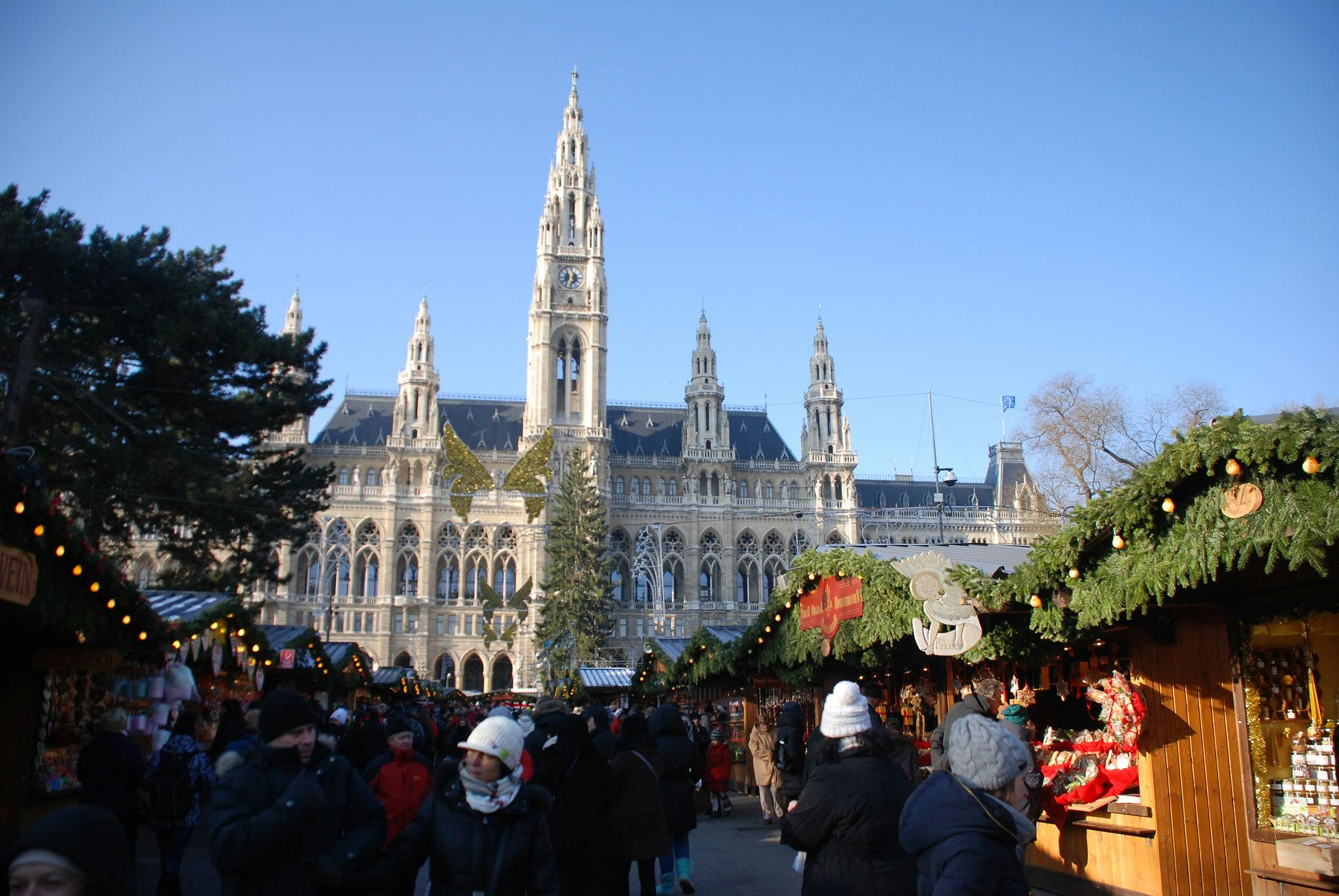 Människor som utforskar festliga marknadsstånd med semesterdekorationer, under en klarblå himmel, framför en storslagen historisk byggnad.